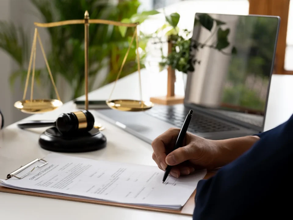 A person signs a legal document. In the background, a scale of justice sits on the desk.