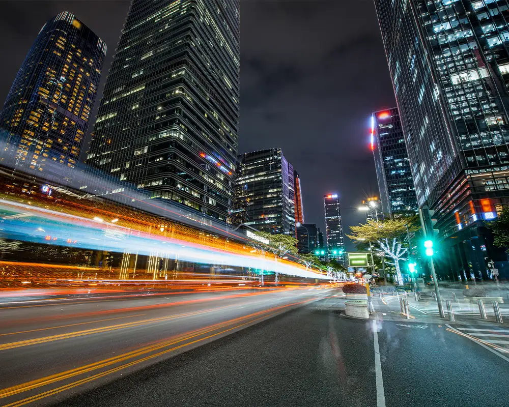 A cityscape at night. Long-exposure vehicle light trails illustrate velocity.