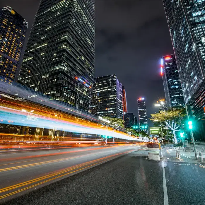 Lights trails streak down a city street at night. Skyscrapers fill the frame.